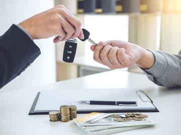 Eine Hand tauscht Autoschlüssel gegen Geld aus; Münzen und Bargeld liegen auf einem Tisch neben einem Klemmbrett und einem Stift, was eine Transaktion symbolisiert.